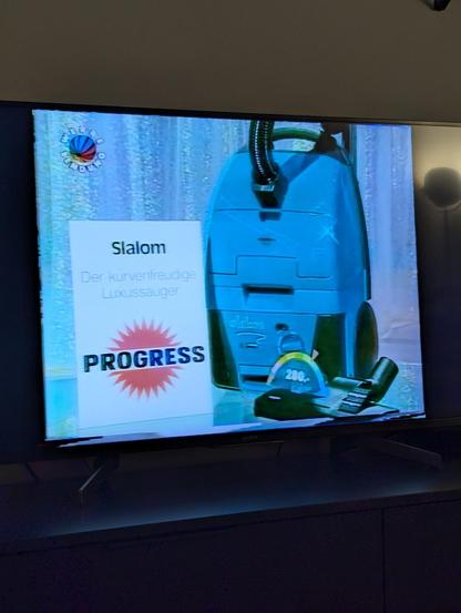 Ein Foto von unserem Fernseher. Darauf sieht man eine Folge Glücksrad aus dem Jahr 1994. Im Bild ist ein blauer Staubsauger mit einem Schild daneben. Darauf steht "Slalom-Der kurvenfreudige Luxussauger von Progress".
