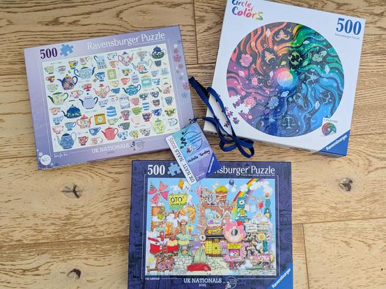 Three jigsaw puzzle boxes laid out on the floor, with my lanyard from UK Nationals lying on top. One is a collection of colourful teapots on a white background, one is a busy but very well-illustrated crazy harbour scene, and the third is a circle puzzle with signs of the zodiac in a gradient of colours.