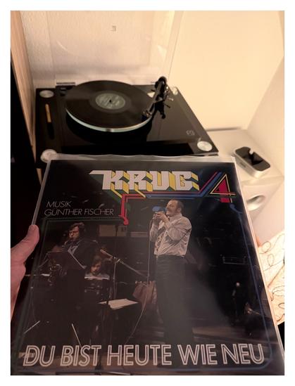 Auf einem schwarzen Plattenspieler läuft eine schwarze Schallplatte. Davor halte ich das Cover ins Bild. Foto, eine Bigband auf einer Bühne. Vorne steht Manfred Krug, Jeans, reingestecktes Hemd, viel zu kurze Krawatte, dafür aber mächtig dicke Koteletten und ordentlich Schnörres. Er singt gerade leidenschaftlich und hält das Mikrofon wie eine Trompete vor den Mund. Der Titel der Platte ist in 3D Schrift. Links steht noch Musik: Günther Fischer.