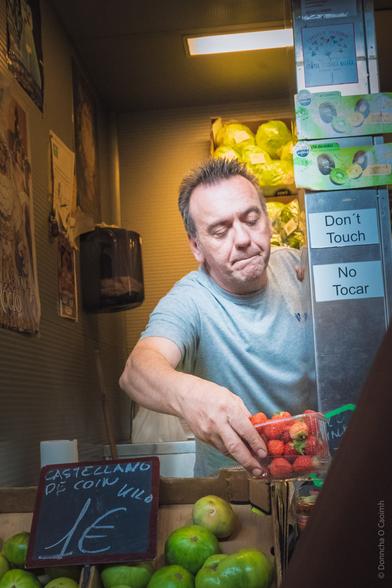 A man in a grey T-shirt is arranging a punnet of strawberries at a market stall in Malaga. Green apples are displayed in the foreground with a sign reading "CASTELLANO DE COIN 1€/kilo." Behind him, there are shelves of lettuce. Signs on the stall read "Don't Touch" and "No Tocar."