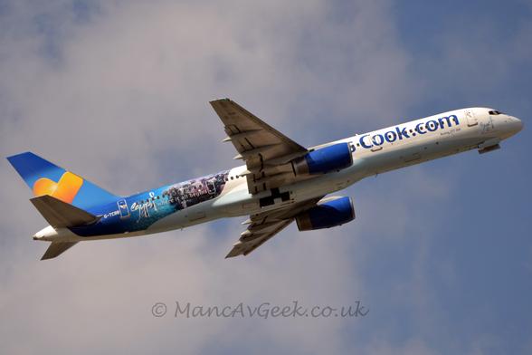 Side view of a white, twin engined jet airliner with a pale blue belly, flying from left to right with the undercarriage doors in the middle of closing, flaps extended from the rear of the wings, and the nose raised significantly, suggesting it has just taken off.
There are blue "Thomas Cook[dot]com" titles on the forward fuselage, partially obscured by the wings and blue engine pods.
The rear fuselage is covered in an image of clear blue sea and grey stone buildings, overlaid with the words "Egypt where it all begins" and the registration "G-TCBB" in white.
The tail is dark and light blue, with a yellow and orange love heart in the middle.
Pale blue sky and fluffy grey and white cloud fill the rest of the frame.