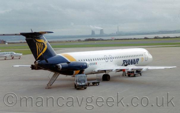Side view of a white, twin engined jet airliner with the engines mounted on the sides of the rear fuselage, parked facing to the right and slightly away from the camera.
There are dark blue "RyanAir" titles on the lower forward fuselage next to a yellow, winged Irish harp.
A larger version of the harp is on the dark blue tail/
The rear fuselage is the same dark blue, with a diagonal yellow stripe in front, with the dark blue engine pods mounted on the sides.
The registration "EI-CCU" is on the side of the rear fuselage, partially obscured by the engine pod.A luggage cart with a couple of trailers is parked in front of the plane, with only a couple of cases on the rear trolley.Grey concrete apron fills most of the frame, with a white twin engined BizProp parked on the left edge of the frame.
Grass in the background leads up to a wide river in the distance, with an industrial landscape on the far side, under grey sky.
