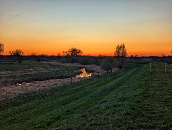 A tranquil, wide shot captures a rural landscape at sunset. The sky transitions from a pale blue at the top to a vibrant orange and yellow along the horizon. Silhouetted trees with bare branches line the distant horizon.
A small, winding stream reflects the warm colours of the sky as it meanders through a field of dry, beige grasses and reeds. The foreground is dominated by a grassy embankment, sloping upwards from left to right. Three white posts with red bands are positioned along the crest of this embankment on the right side of the frame. The overall mood is peaceful and serene, typical of a countryside evening.
