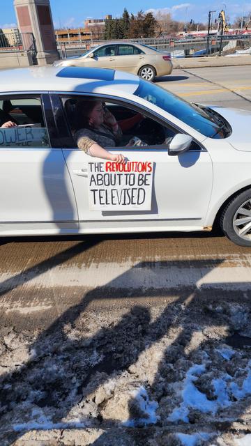 The image shows a white car on a road with a person inside holding a sign. The sign reads "THE REVOLUTION'S ABOUT TO BE TELEVISED" with "REVOLUTION'S" in red and the rest in black. The person is wearing a patterned top and has their arm extended out of the car window. The car is parked on the side of the road, and there is a visible shadow on the ground, indicating a sunny day. In the background, there is another car and a bridge, with some construction equipment visible. The road appears to be partially cleared of snow, suggesting a winter season. The sky is clear with a few clouds, and the overall scene suggests a protest or demonstration.