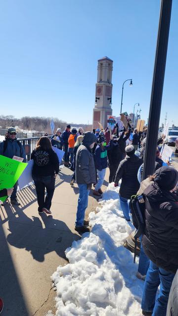 The image depicts a group of people gathered on a bridge during a sunny day, with snow visible on the ground, indicating a cold season. The participants are holding various signs, some of which are visible, including one that reads "WE HAVE NO CHOICE" in bright green. The crowd is diverse, with individuals wearing winter clothing such as jackets, hats, and gloves. The bridge features a tall clock tower in the background, and the sky is clear and blue. The scene suggests a protest or demonstration, with people walking and standing, some raising their signs. The overall atmosphere is one of organized activity, with participants engaged in a collective cause.