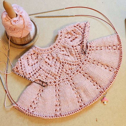 A pink knit dress in progress. The top has a lace triangle pattern with the skirt in small sections with a strip of super basic lace. There is a small mushroom stitch marker, with a pink top and little eyes and a smile on the stalk.