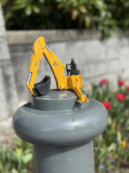 Part of a yellow and black toy digger sitting on top of a grey bollard with some flowers and a grey wall in the background. The toy part includes the scoop arm and bucket. It looks like it was placed there in case a searching parent is looking for it. A parent is almost certainly searching for it.