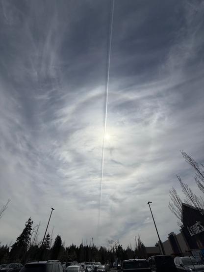 Wide lens picture of the sky over a Lowe’s parking lot. The sun is bright through streaky thin cloud cover in a blue sky; bright enough to create a sort of halo effect around the sun. An airplane contrail straight through the sky appears to have a shadow on the clouds. Some of the weirdness may be iPhone camera madness. It looks cool though.