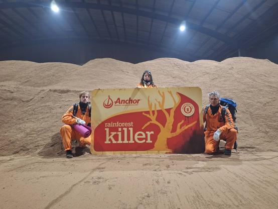 Three people with an oversized butter block sit on pile of palm kernel in a shed.
The butter block says Anchor Rainforest Killer.