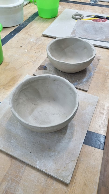 Two slightly misshapen unglazed and unfired 25cm clay noodle pots on wooden boards on a plywood bench in a pottery studio.