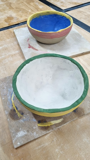 Two slightly misshapen 25cm clay noodle pots, painted, unglazed and as yet not fired on wooden boards on a plywood bench in a pottery studio.