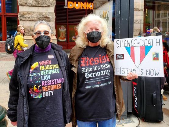 Two women standing at the Hands Off Rally. Their shirts and sign are printed with the slogans in the post.