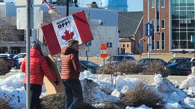 The image depicts two individuals walking on a snow-covered sidewalk in a parking lot area. The person on the left is wearing a bright red jacket and a knit hat, carrying a cardboard sign. The person on the right is wearing a brown jacket and a green beanie, holding a large Canadian flag with the text "HANDS OFF!" prominently displayed. The background features a mix of buildings, including a modern glass-fronted structure and a brick building, with parked cars and a clear blue sky. A road sign indicating "CITY WEST" is visible, along with a Canadian flag on a pole. The scene suggests a protest or demonstration, with the Canadian flag and the "HANDS OFF!" message indicating a political or social statement.