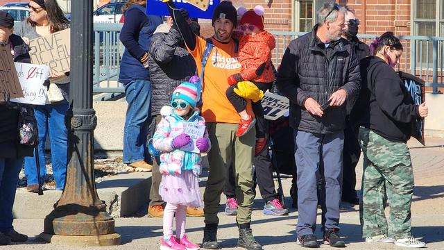 The image depicts a group of people gathered outdoors, likely participating in a protest or demonstration. In the foreground, a young girl wearing a colorful winter outfit, including a pink tutu, a multicolored jacket, and a hat with pom-poms, is holding a sign. She is accompanied by an adult male in an orange shirt with the text "This Guy Cares" and green pants, who is holding a child dressed in a red outfit with a hood. The background shows several other individuals, some holding signs, including one that reads "SAVE OUR CHILDREN." The setting appears to be a public space with a brick building in the background, and the atmosphere suggests a peaceful gathering.