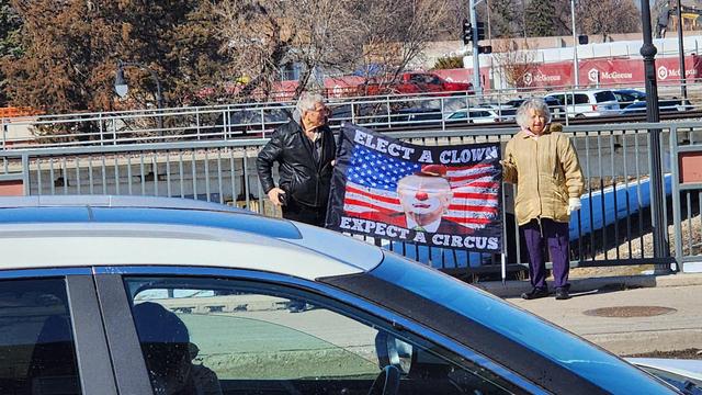 The image shows two individuals standing on a sidewalk next to a metal railing, holding a large banner. The banner features an image of a clown with a red nose and a white face, set against a background of the American flag. The text on the banner reads "ELECT A CLOWN EXPECT A CIRCUS." The individuals are dressed in winter clothing, with one wearing a black leather jacket and the other in a tan coat. In the foreground, a car is visible, partially obscuring the view. The background includes a bridge with vehicles and a construction site with a McDonald's sign. The scene appears to be set in a public area, possibly during a political event or protest.