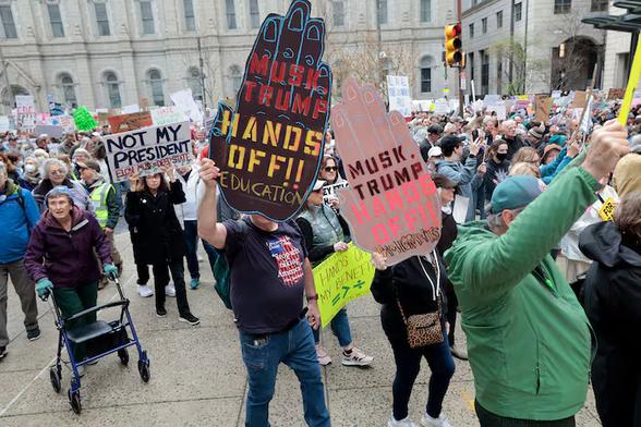 Photo of the Hands Off rally in Philadelphia, Saturday 5 April, by Elizabeth Robertson for the Philadelphia Inquirer. A large crowd on the north side of Philadelphia City Hall. One prominent sign reads "NOT MY PRESIDENT - ELON MUSK"; two people are carrying three-feet tall hand-shaped signs, saying "MUSK-TRUMP HANDS OFF" Education and Immigrants. The crowd also contains people using mobility assistive devices such as walkers and crutches.