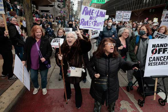 Photo of the HandsOff rally in Philadelphia, Saturday 5 April, by Elizabeth Robertson for the Philadelphia Inquirer. As the crowd moves from City Hall to Independence Hall, signs saying "HANDS OFF CANCER RESEARCH" and "RULE OF LAW - NOT RULE OF TRUMP" are prominent. Some people are wearing N95-style masks; some are sing assistive devices such as walkers and canes. Other folks are standing on the sidewalk along the route, cheering and carrying their own signs.
