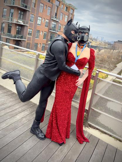 A fun moment between two human pups in a playful pose - one wearing a sleek black leather outfit complete with a detailed leather puppy hood featuring pointed ears and a nose ring, while the other wears a stunning floor-length sparkling red gown with long sleeves and a matching leather puppy mask. The figure in black has one leg playfully lifted behind them as they lean into the other pup, creating a romantic silhouette against the backdrop of brick apartment buildings and distant city skyline. The red-dressed figure wears a blue lanyard, possibly an event credential, with a yellow pendant. Both are positioned on wooden decking with metal railings, with patches of early spring vegetation visible in the background. This intimate moment takes place during the International Puppy & Trainer Contest (IPTC) weekend in Chicago, which is currently happening April 3-6, 2025 at the Center on Halsted.