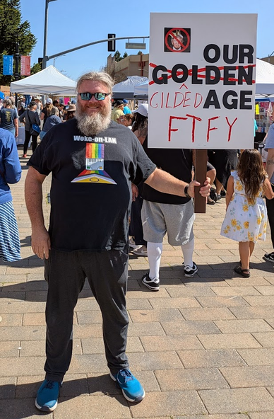 The author wearing a t-shirt with the text "Woke-on-LAN" which features an Ethernet port with the New Progress Pride flag passing through and behind the port. He is a heavyset white man with a full, greying beard wearing reflective sunglasses, charcoal pants and blue sneakers. He is holding a protest sign which has a photo of Donald Trump wearing a crown with a circle and slash superimposed over his ugly face, and text that reads "Our Golden Age" but "golden" is crossed out with red marker and a red carat pointing to the crossed out word, beneath which is the word "gilded." At the bottom of the sign the same red marker reads "FTFY." In the background are numerous protesters and pop-up tents.