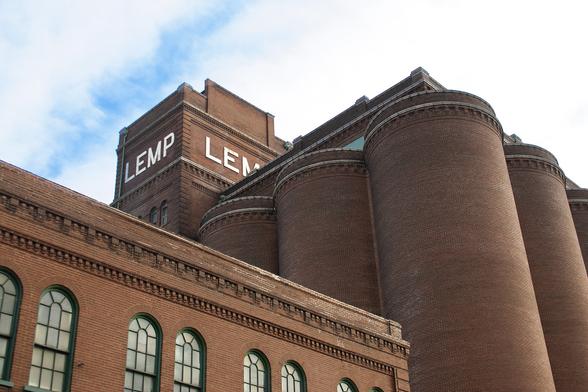 A color photograph of part of the Lemp Brewery complex, in St. Louis, Missouri USA. The building is red brick, with a cluster of cylindrical structures at the right of the frame and a shorter wing with arched windws at the left of the frame. near the center, as rectangular tower with the word "LEMP" in white, sans-serif capitals rises above.