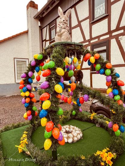 Der geschmückte Osterbrunnen in Münster aus der Frontalperspektive: Bunte Ostereier hängen an grünem Girlandenschmuck in Glockenform. In der Mitte des Brunnens steht eine Hasenfigur, dahinter ein Fachwerkhaus.