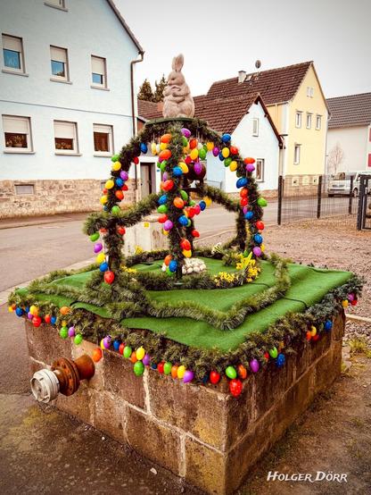Seitliche Ansicht des Osterbrunnens: Die grüne „Osterglocke“ mit bunten Eiern spannt sich über den Brunnen. Im Vordergrund blühen gelbe Narzissen, im Hintergrund sind Häuser sichtbar.