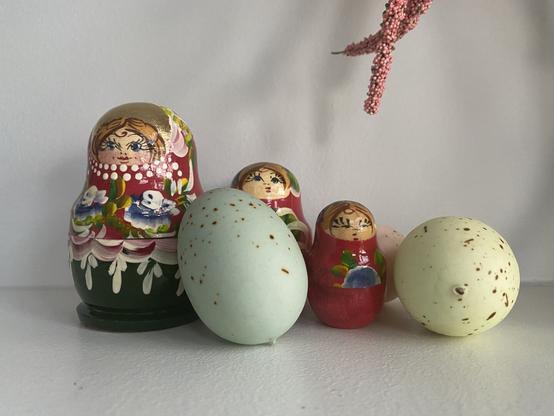 3 red and gold stacking matryoshka dolls a s two faux speckled eggs, blue and green pastel, on a white table.