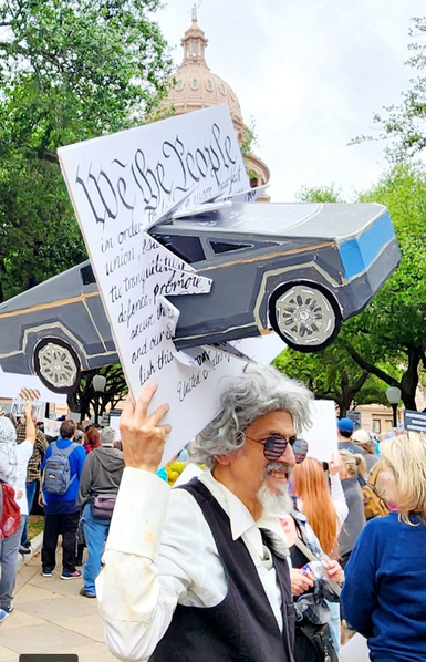 Gentleman in a suit wearing American flag sunglasses holding a cardboard sign with the intro to the Constitution and a 3D cybertruck plowing through the middle of it.