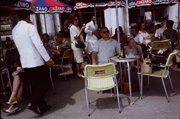 A man with a military crew cut wears dark tinted glasses. He's leaning back in a cafe chair. To his left is a boy about ten who has a similar hair cut and is completely asleep in his chair.  Waiters in white suit jackets and dark trousers serve various guests. Each table has a large blue and red umbrella with the CinZano logo. The backs of the chairs do too.