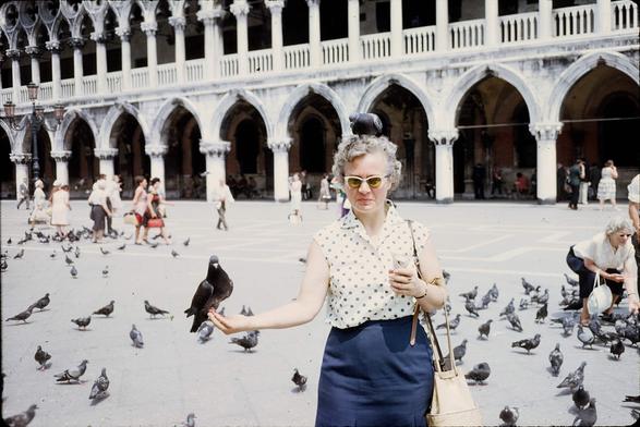 A white haired woman in horn rimmed sunglasses holds her right hand outstretched. A pigeon is in her hand. There's another on her head. Behind her is the arcade of Piazza San Marco, as well as dozens of other pigeons and other tourists who are more interested in the birds than the building.
