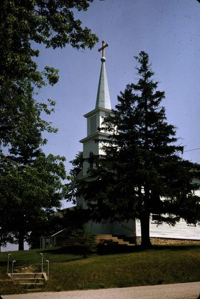 A classic white washed American church with a single steeple that's just visible among the pine trees. A path and stairs through the lawn lead to the sidewalk and street.