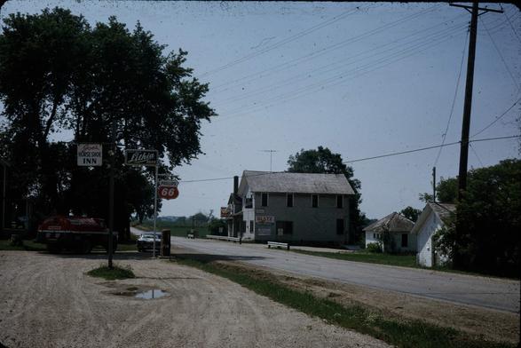 A country road with a gravel paved Philips 66 service station with a sign for Joe's Horseshoe Inn. Further down the road is a two story wood building that offers food. Currently it's a restaurant and might have been back then.