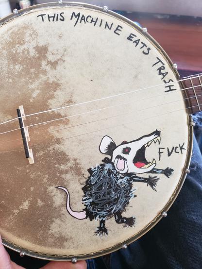A banjo with a scribbly possum shouting "fuck" and the phrase "this machine eats trash" painted on it