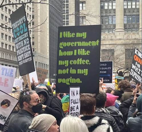 Photo of a protest march focused on a sign that says "I like my government how I like my coffee, not in my vagina."