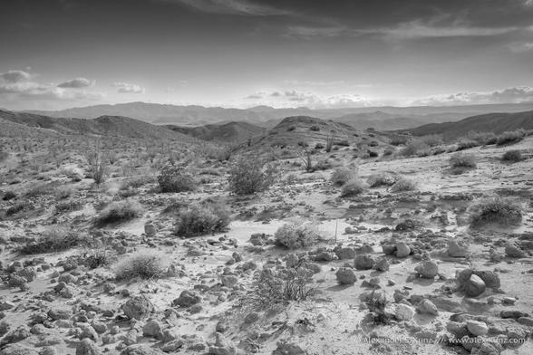 Black & white photo of a desert landscape. Rocks are strewn about a gently sloping sandstone plateau with hardy shrubs. Low clouds hang above a mountain range in the distance, decorating an otherwise mostly clear sky.