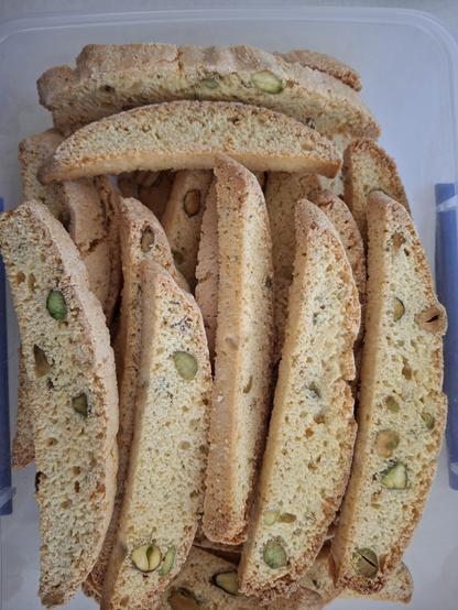 A container full of long narrow, golden brown strips of baked biscotti. There are pistachios dotted throughout the biscuits.