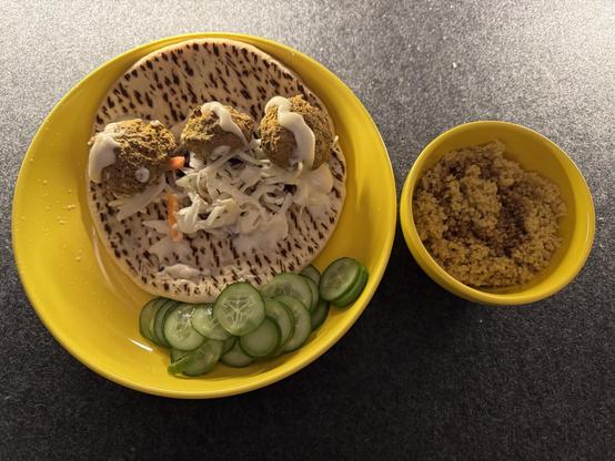 Moroccan dish with falafel, diced cabbage, and garlic mayo on a pita in one dish. Sliced cucumbers sit next to the pita. In another small cup is Moroccan couscous.