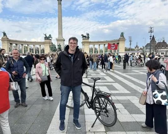 Ein belebter Platz mit vielen Menschen und Fahrrädern. Im Vordergrund steht ein lächelnder Mann mit dunkler Jacke neben einem Fahrrad. Im Hintergrund sieht man ein großes, halbkreisförmiges Denkmal mit Reiterstatuen und einer hohen Säule. Eine rote Fahne mit einem Antifa-Symbol ist in der Menge zu sehen. Der Himmel ist teils bewölkt.
