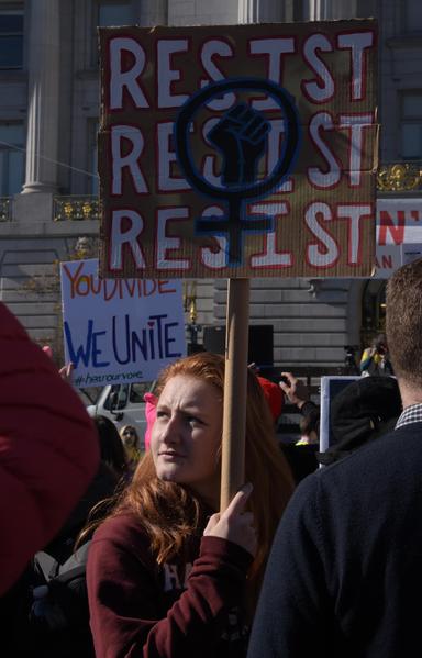 a woman holds a sign that says 
RESIST
RESIST
RESIST
with a fist and the woman symbol