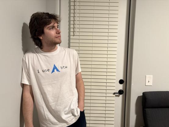 A young man indoors leans against a wall wearing a white t-shirt. The t-shirt sports the Arch Linux project logo, and the text "i use" to the logo's left, and "btw" to the logo's right.