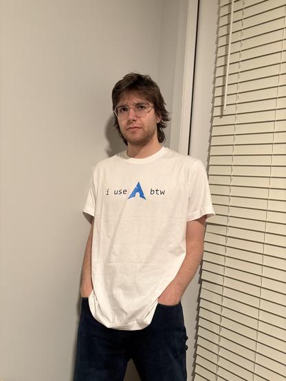 A young man indoors leans against a corner wearing a white t-shirt. He is wearing geometric prescription glasses and is looking into the camera. The t-shirt sports the Arch Linux project logo, and the text "i use" to the logo's left, and "btw" to the logo's right.
