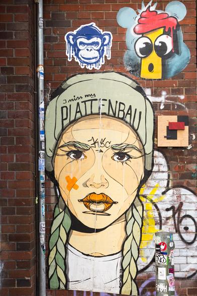 Picture of a young woman, pasted to a brick wall;
the woman wears a cap with the words "I miss my PLATTENBAU"
