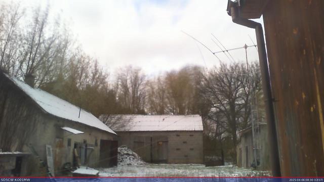 A webcam view of the SO5CW antennas: A triband yagi on a small tower and a vertical antenna (that used to be more straight) in the back of the yard. A barn and an old auxillary building are visible around the yard. There is a little snow on the ground and the roofs, the sky is covered in clouds.