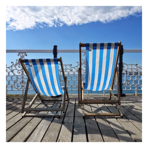 This is a square format colour image of two empty decks chairs on a pier. They are positioned away from the viewer, looking out towards the sea and Brighton beach. Backlit, the blue and white vertical stripes really pop together with the blue sky and white clouds. This is an image of lazy summer days by the sea. The wooden slats of the pier lead the viewer towards the chairs and beyond. Along the middle of the frame, behind the chairs, is the ornate metalwork of the Victorian pier. Taken in Brighton with a Sony RX100.