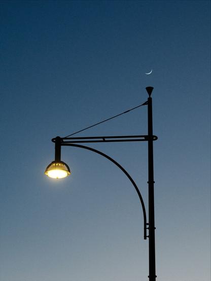 Das Foto zeigt eine Straßenlaterne vor einem wolkenlosen Himmel. Über ihr erscheint die schmale Sichel des zunehmenden Mondes.

The photo shows a street lamp against a cloudless sky. The narrow crescent of the waxing moon appears above it.
