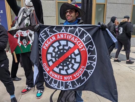 Un manifestant jovial, chapeau de sorcière sur la tête, tient un drapeau : Païen⋅ne⋅s anti-fascistes, nos runes pas les vôtres.