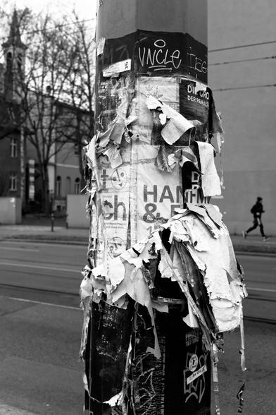 Der untere Teil eines Laternenmastes ist mit Aushängen beklebt. Von diesen sind mehrere Schichten übereinander zusehen.