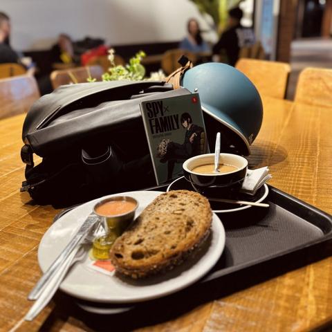 Bodegón de desayuno en mesa comunal. Hay una bandeja con una tostada de tomate y aceite con pan multicereal y un café rico con leche fría de avena. Detrás están el manga que me estoy leyendo (Spy x Family 5), la sudadera, la mochila y el casco de la bici.