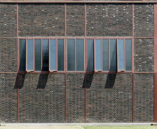 Mur de briques sombres quadrillé de renforts en métal rouge. Au centre, une rangée de fenêtres industrielles rectangulaires étroites dont les montants sont également en métal rouge. Certaines sont ouvertes et projettent une ombre sur le bas du mur.