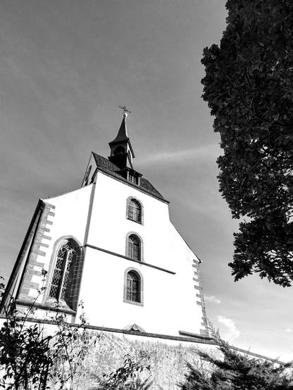 A black and white photograph of a church viewed from a low angle. The church features a tall steeple with a cross at the top, multiple arched windows, and a simple facade. There is a tree partially visible on the right side of the image, and the sky appears clear with some wispy clouds.